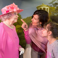 En kvinna och en tjej beundrar en vaxfigur av en äldre kvinna i en rosa outfit och hatt, i en parkliknande bakgrund.