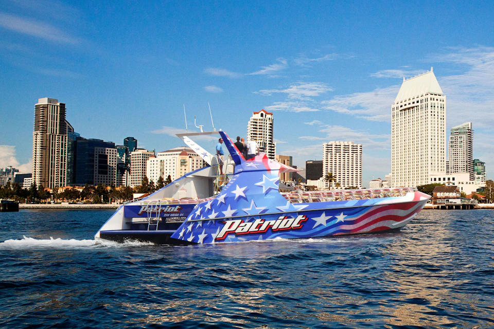 Boat decorated with American flag pattern and "Patriot" name, sailing on water with a city skyline in the background under a clear sky.