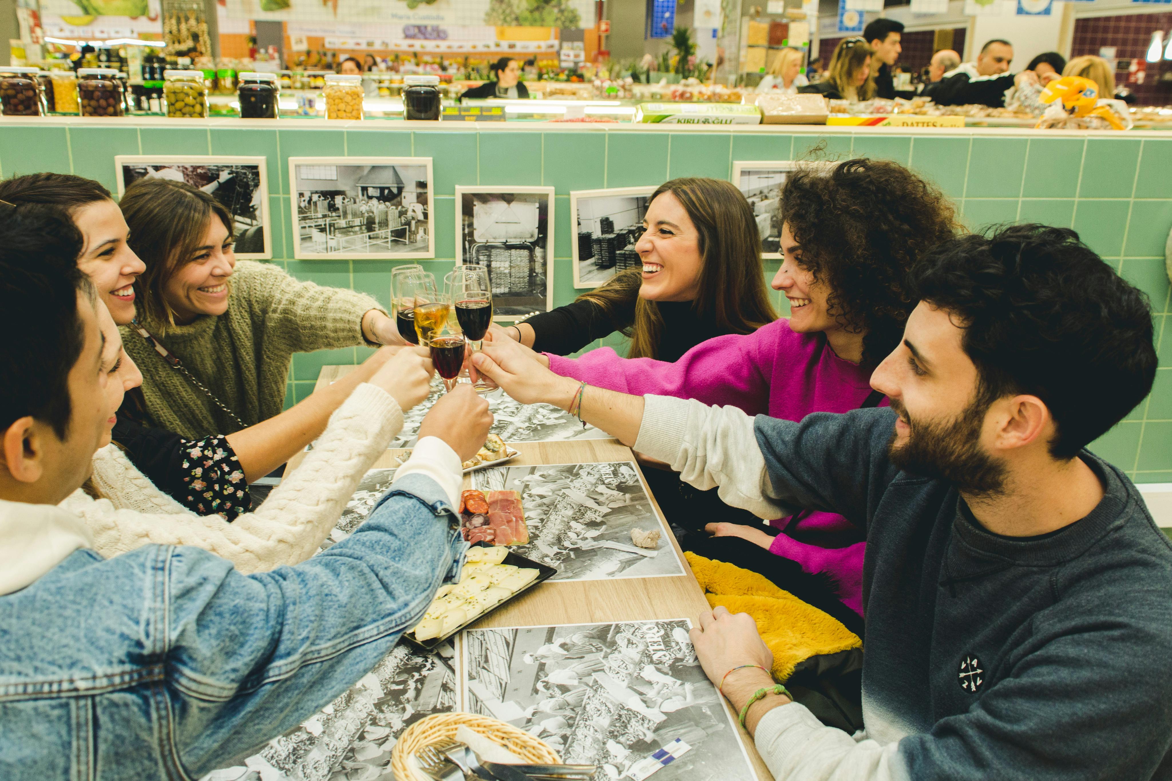 Μια ομάδα έξι φίλων κάνει πρόποση με ποτά γύρω από ένα τραπέζι με φαγητό σε μια ζωντανή, έντονα φωτισμένη εσωτερική αγορά.