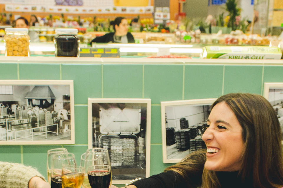 A group of six friends toasting with drinks around a table with food in a lively, brightly lit indoor market.
