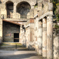 Forntida romerska ruiner med pelare, dekorativa väggar och trappor i en utomhusarkeologisk plats. Övergivna strukturer i bakgrunden.