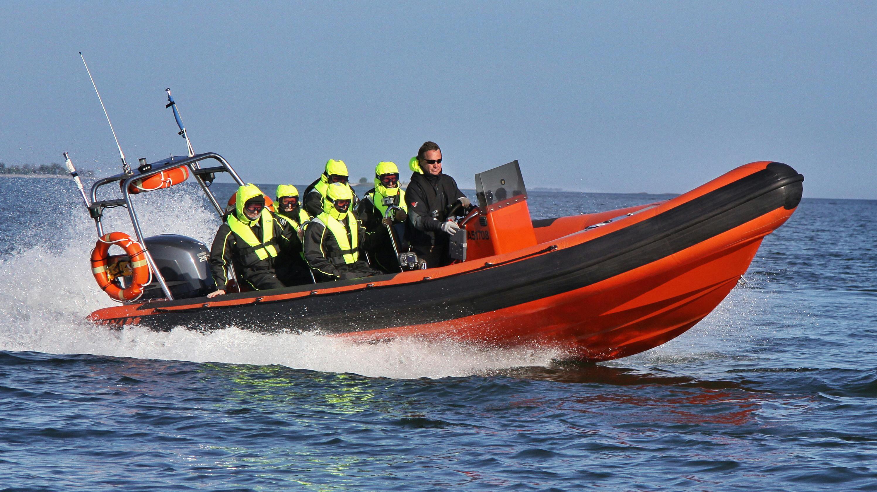 Μια ομάδα ανθρώπων με μαύρα κοστούμια και σωσίβια από νέον καβαλικεύουν ένα πορτοκαλί ταχύπλοο στο νερό, οδηγούμενοι από έναν άνδρα με γυαλιά ηλίου.