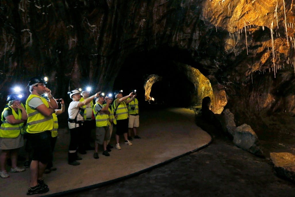 A group of people in safety gear with headlamps and reflective vests explore a dark cave, pointing flashlights at the walls.