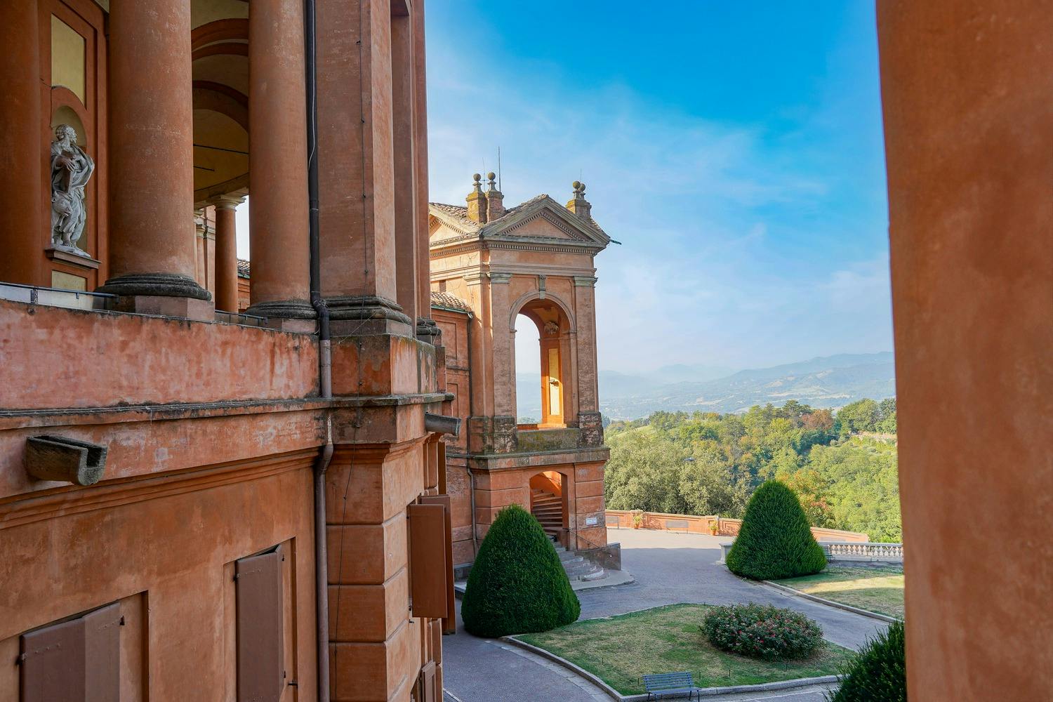 An old, reddish building with arches overlooks a manicured garden and distant, green hills under a clear blue sky.