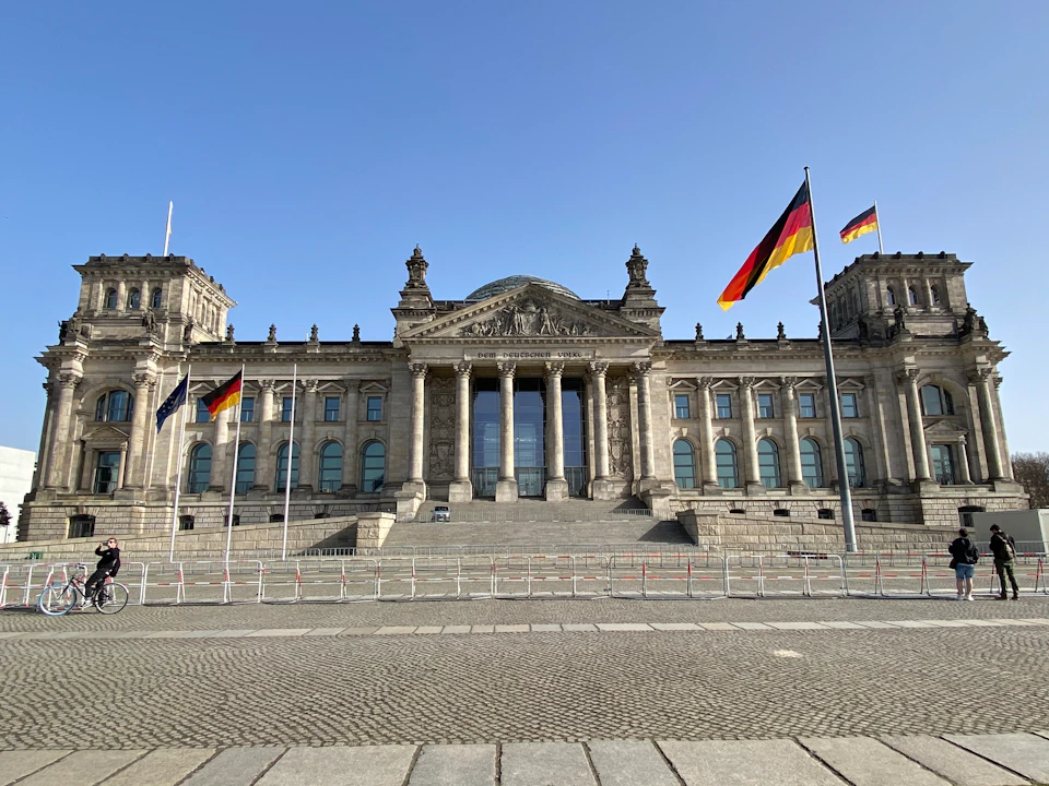 Historic building with columns and a glass dome, flanked by German flags, under a clear blue sky. Cobblestone area in the foreground.