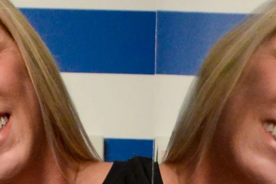 A person in black clothing stands with arms outstretched in front of a striped blue and white background, reflected on a mirror.