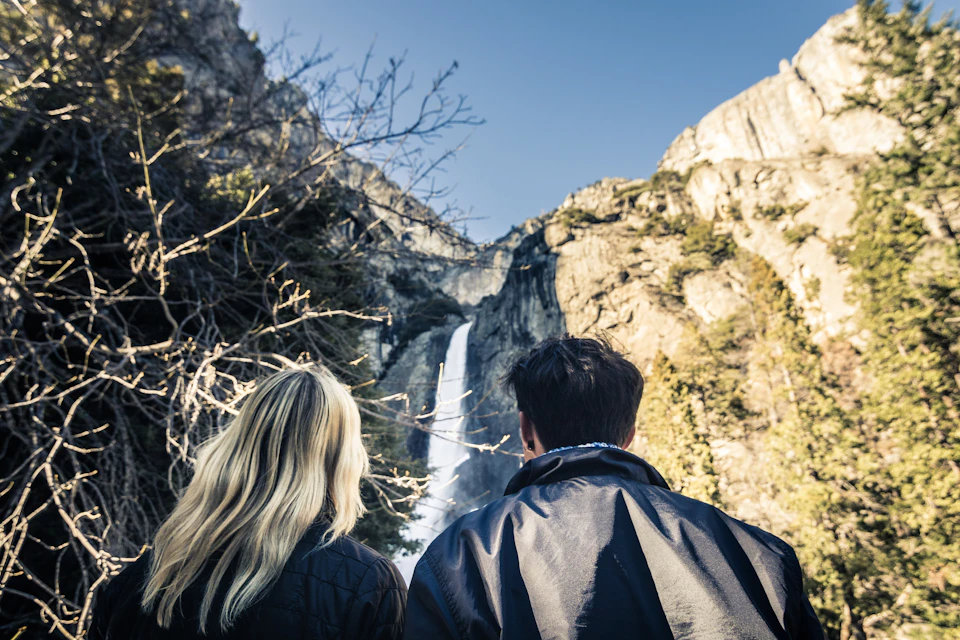 Two people, seen from behind, look at a waterfall flowing from a rocky cliff under a clear blue sky.