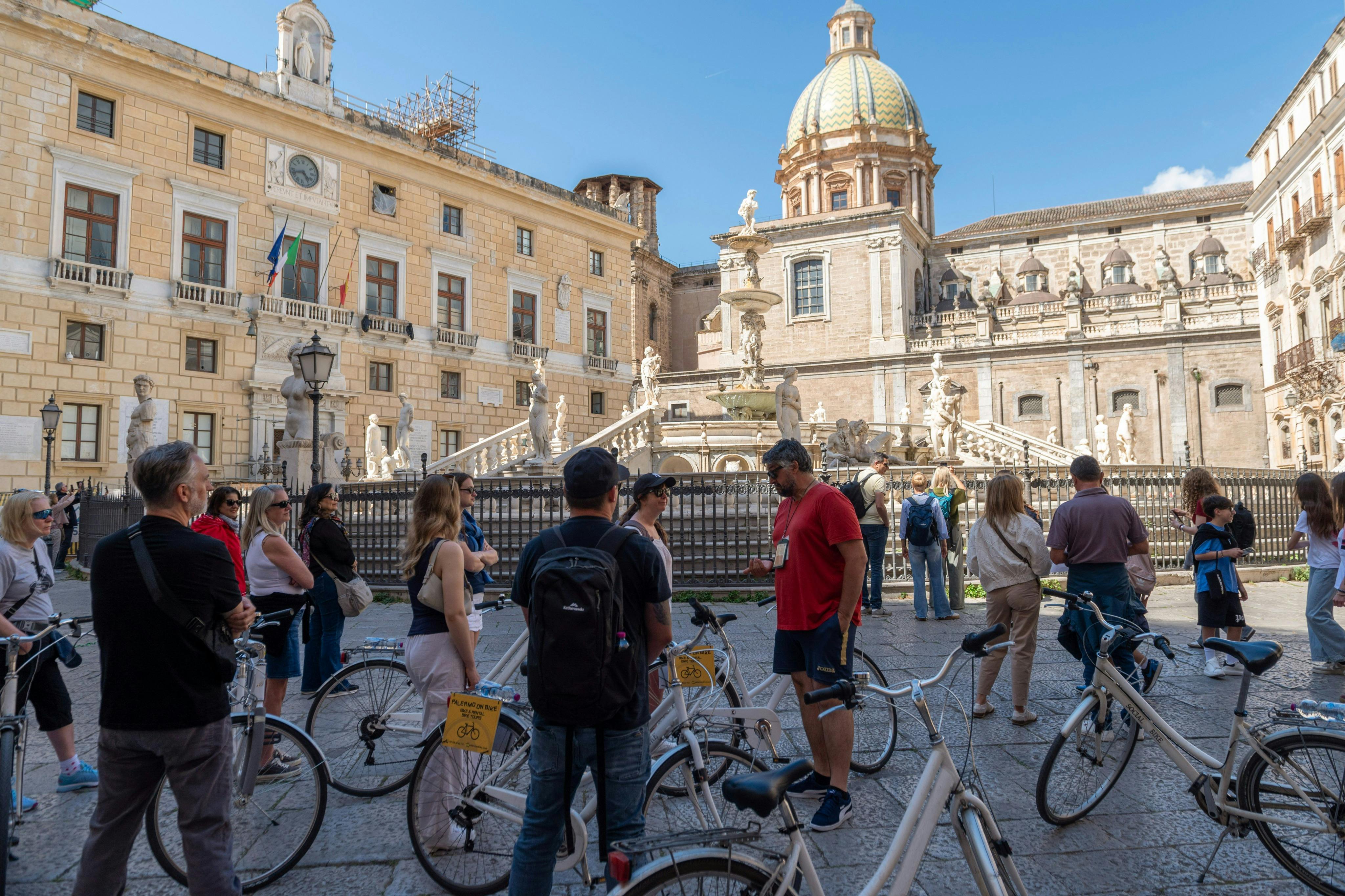 Un groupe de personnes à vélo sur une place historique ornée d'une fontaine et d'un dôme, sous un ciel bleu limpide.