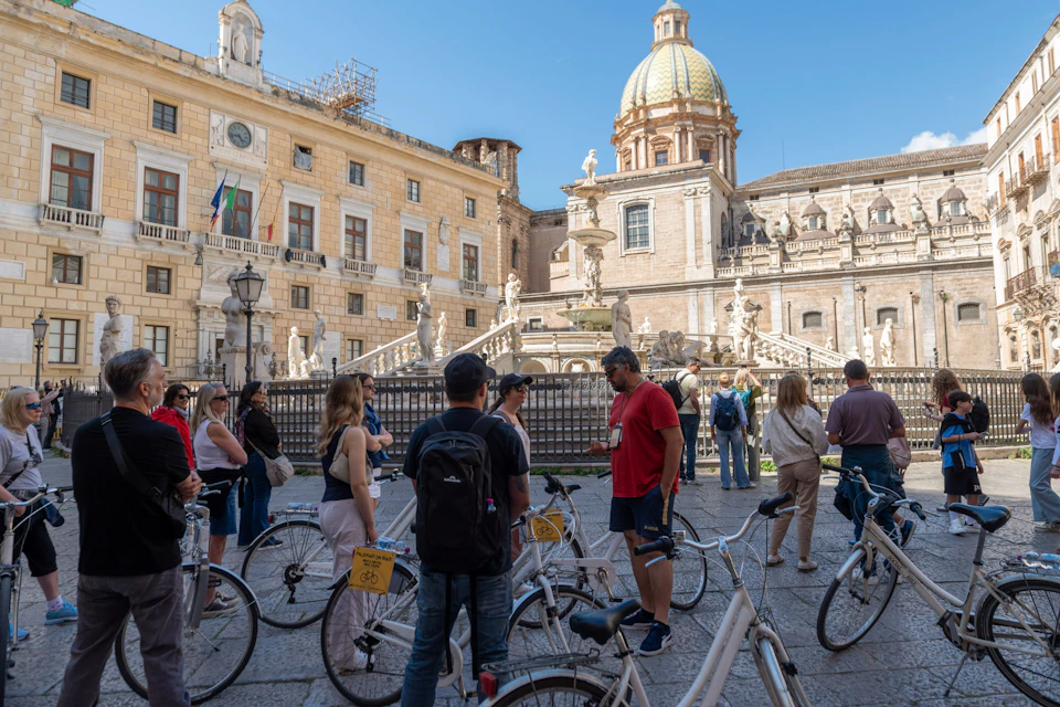 A group of people with bicycles in a historic square featuring a fountain and a dome under a clear blue sky.