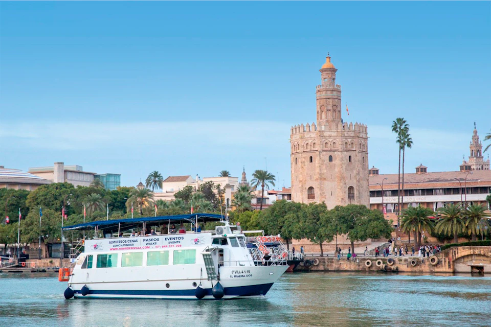A sightseeing boat sails on a river near a historic stone tower surrounded by palm trees and buildings under a clear blue sky.