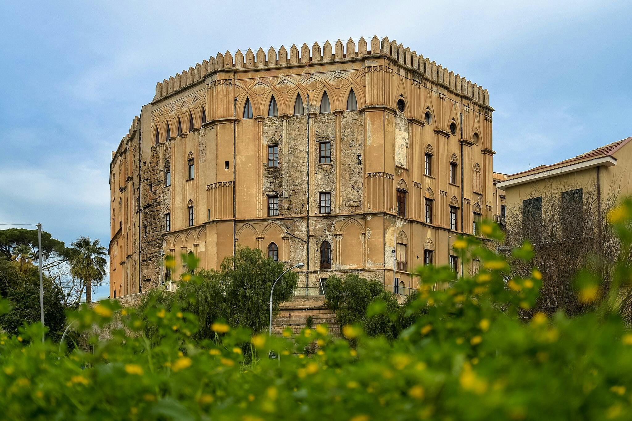 Un gran edificio histórico de color beige con ventanas arqueadas y almenas decorativas, rodeado de vegetación y visto desde un ángulo bajo.