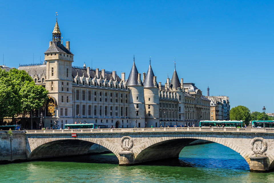 Ornate historical building with conical towers and a bridge over a river, under a clear blue sky.