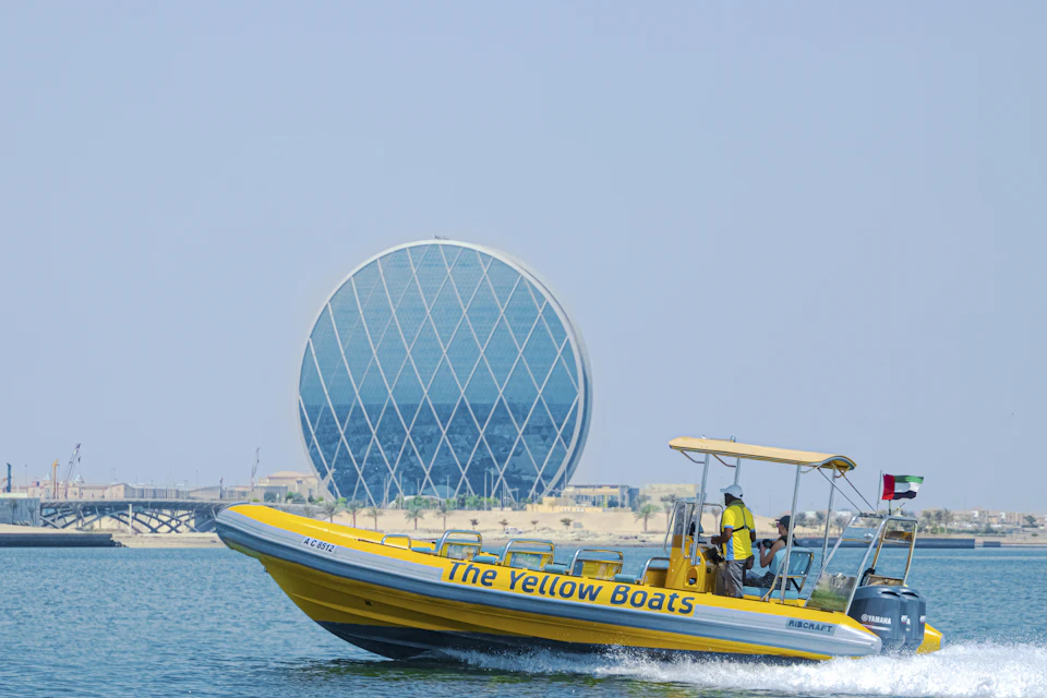 A yellow speedboat labeled "The Yellow Boats" on water with a round, glass-paneled building in the background under a clear sky.