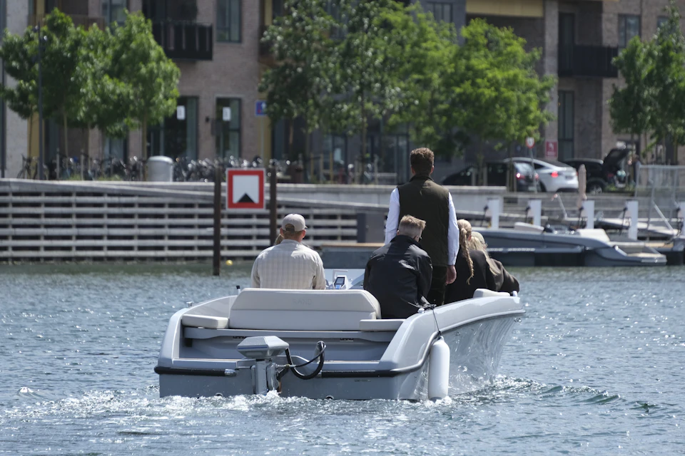 A small group of people riding a motorboat on a calm urban waterway, with buildings and greenery in the background.