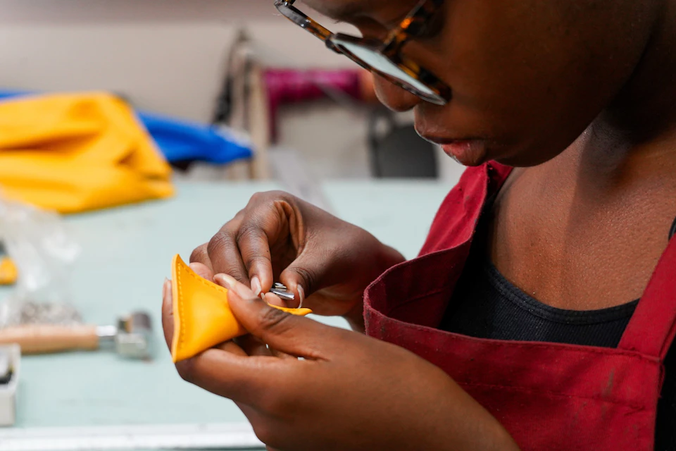 Person sewing a yellow fabric piece, wearing glasses and a red apron, focused on the detail work.