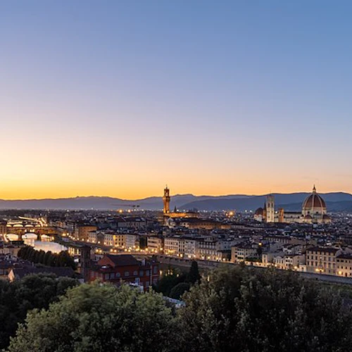 Panoramautsikt över en stadsbild i solnedgången, med historiska byggnader, en flod och avlägsna berg under en lutande himmel.