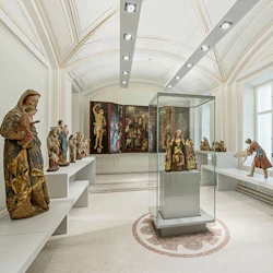 Ett museum som visar religiösa statyer på bänkar och i ett glasfodral, med en stor triptykmålning på bakväggen.