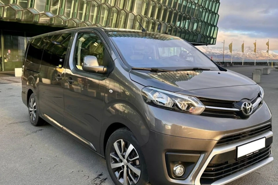 A gray minivan parked outside a modern glass building with hexagonal patterns, under a partly cloudy sky.