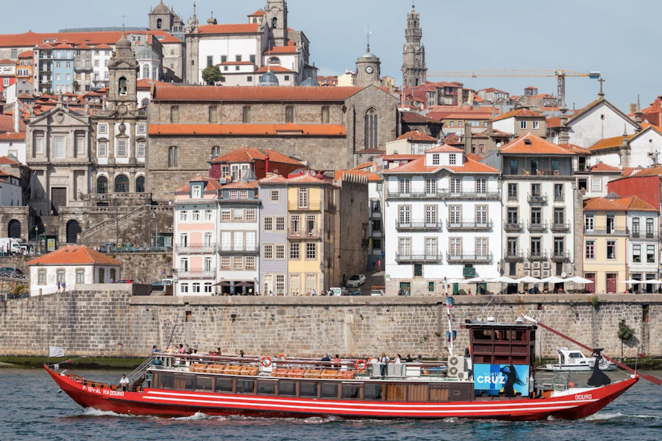 A tour boat passes by colorful, historic buildings along a river in a European city. Towering church structures are visible in the background.