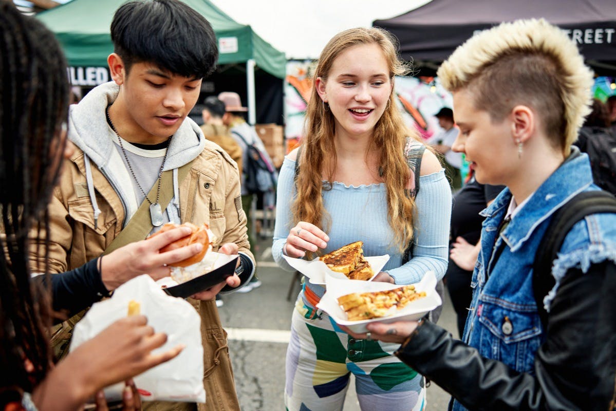 Τρεις νεαροί ενήλικες απολαμβάνουν φαγητό σε εξωτερικούς χώρους σε μια υπαίθρια αγορά, χαμογελώντας και αλληλεπιδρώντας, με πάγκους φαγητού και γκράφιτι στο παρασκήνιο.