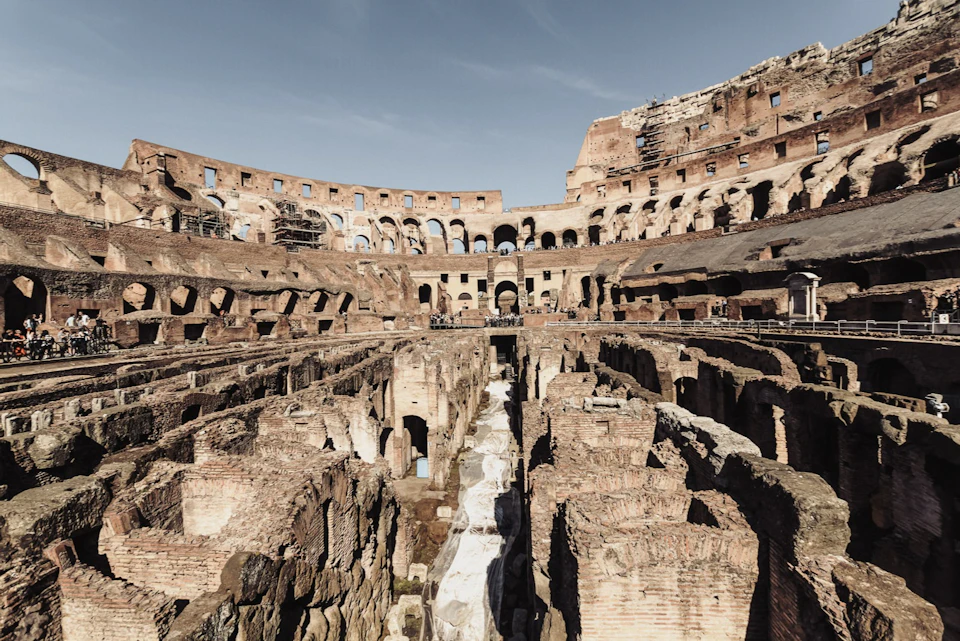 Interior view of the Colosseum in Rome, showing the ancient stone structure and underground passages under a clear sky.