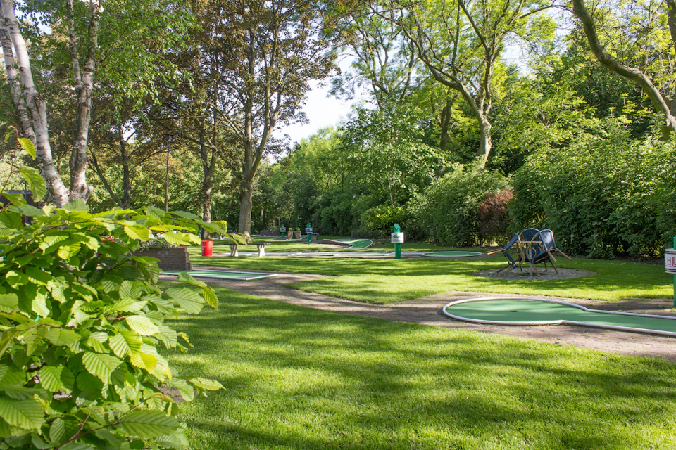 A landscaped mini-golf course with green putting greens, surrounded by trees, grass, and benches in a park setting.