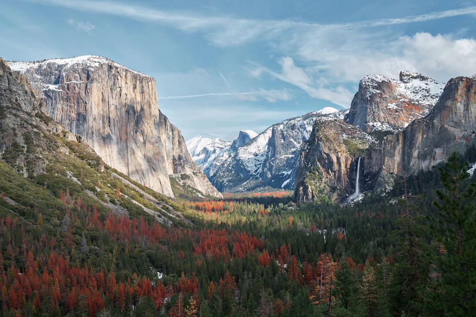 Snow-capped mountains and a waterfall overlooking a forest with green and red trees under a partly cloudy sky.
