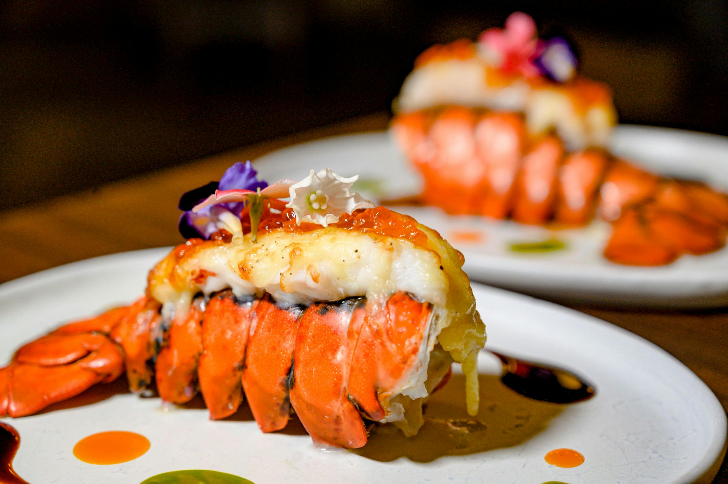 A close-up of a plated lobster tail dish garnished with edible flowers and served with colorful sauces.