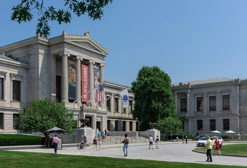 People walk in front of a large, ornate building with columns and banners, including "WELCOME," under a clear blue sky.