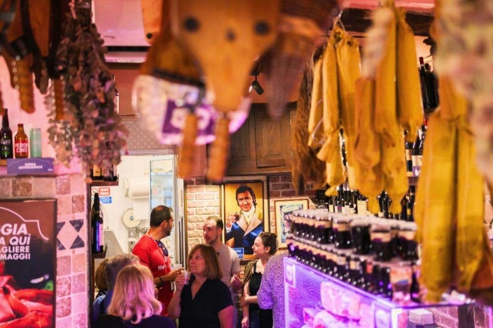 A group of people conversing inside a cozy, decorated room with hanging items and shelves of bottles under warm lighting.