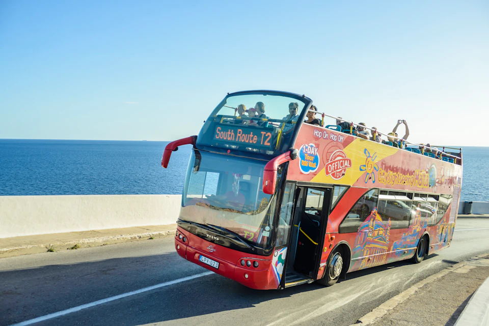 A red double-decker tour bus with open top carrying passengers travels along a coastal road beside a calm blue sea.