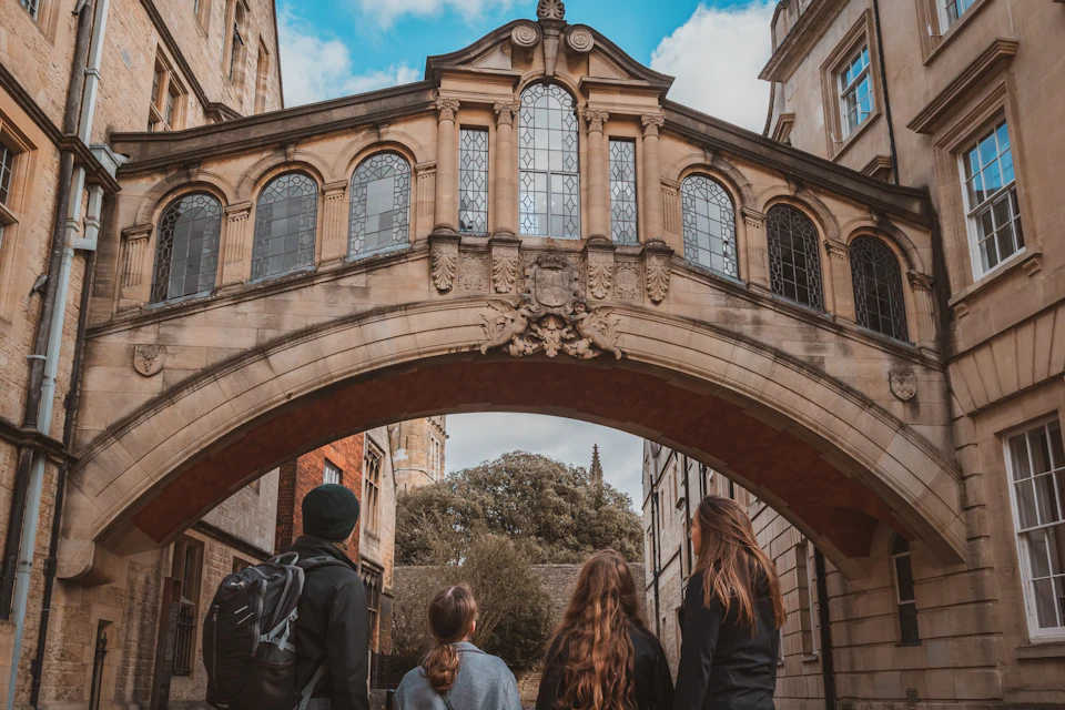 Four people standing under a historic stone bridge with arched windows, surrounded by old buildings and a clear sky.