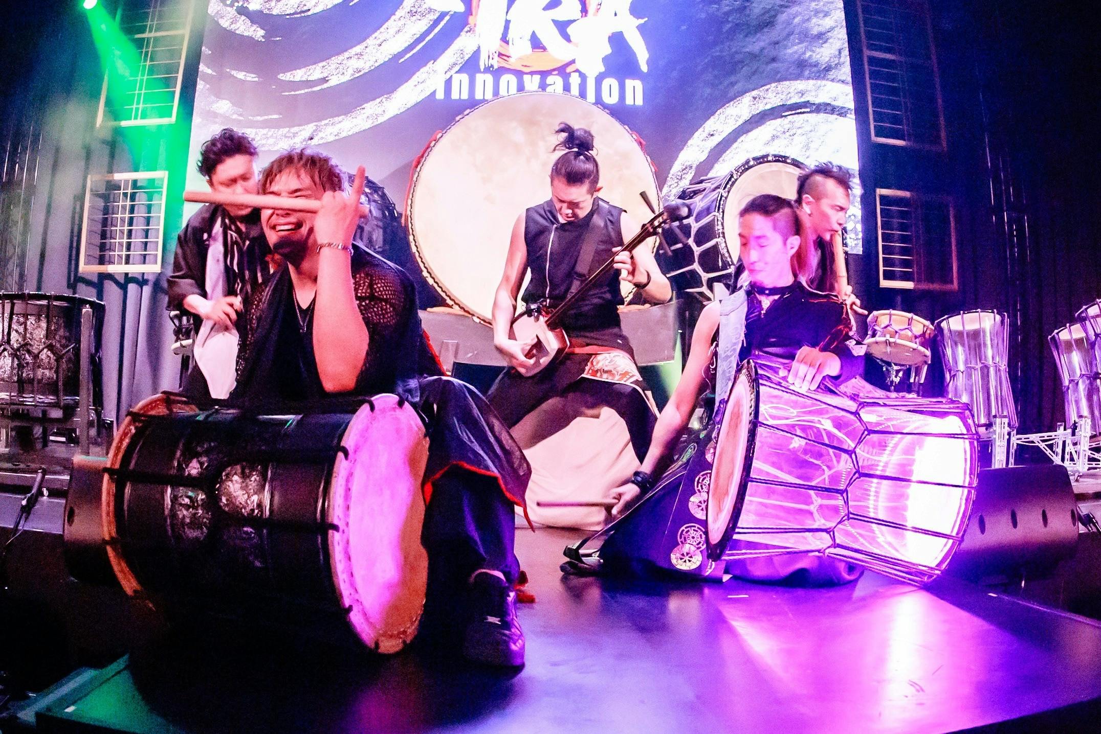 A group of performers playing traditional drums on a stage under colorful lighting, with energy and motion visible.