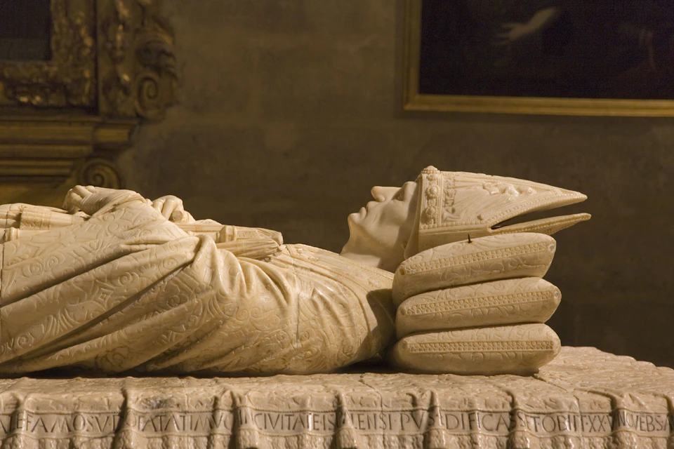 A marble effigy of a medieval figure lies in repose, hands crossed, adorned in detailed robes and a mitre, on a carved stone slab.
