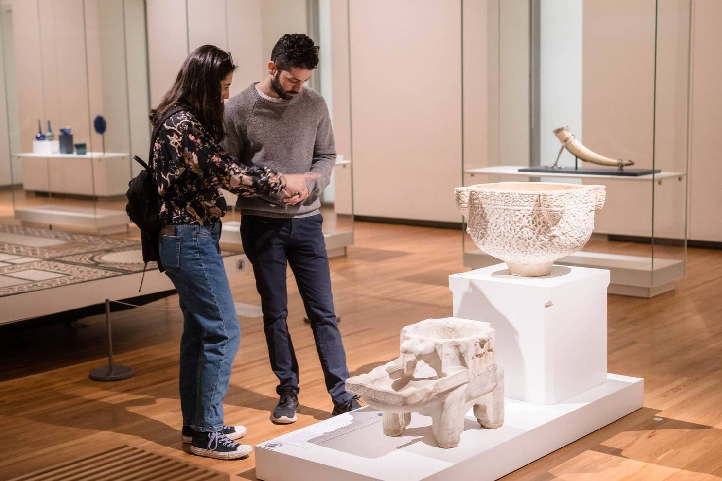 Dva lidé v muzeu se dívají na chytrý telefon. Kolem nich jsou vystaveny různé starověké artefakty.