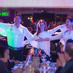 Artister i vita skjortor som dansar på en scen, med sittande publik som klappar i förgrunden.