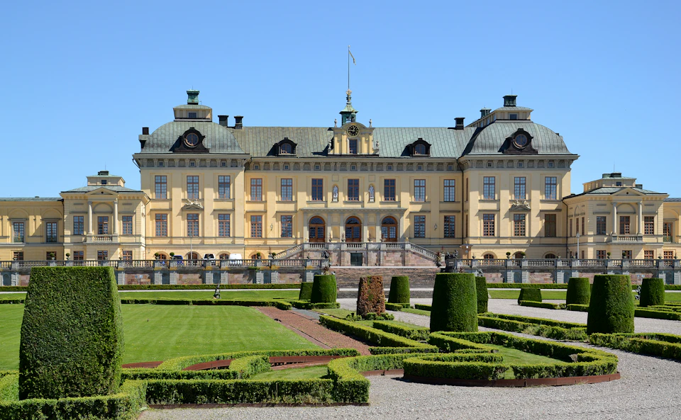 Grand, yellow baroque palace with symmetrical windows, ornate roofs, a central staircase, and manicured gardens in front.