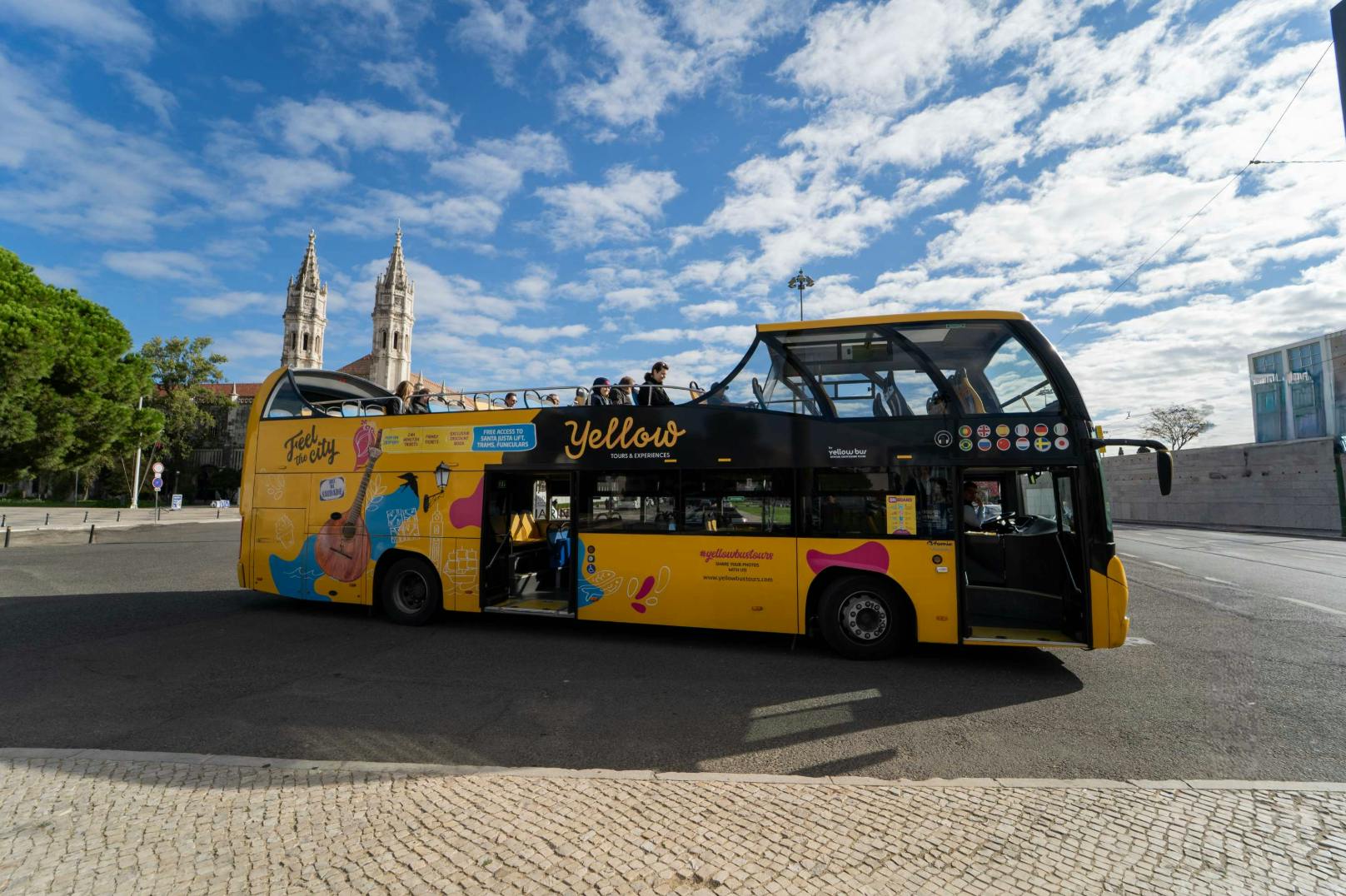 Un autobús turístic groc de dos pisos amb la part superior oberta està aparcat prop d'una plaça, amb una església visible al fons.