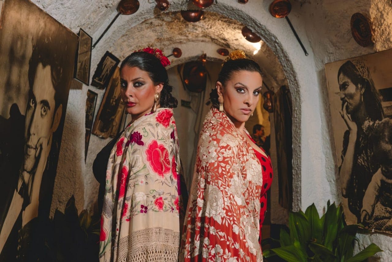 Two women wearing traditional, colorful shawls stand back-to-back in an ornately decorated hallway with copper pots and framed photos.