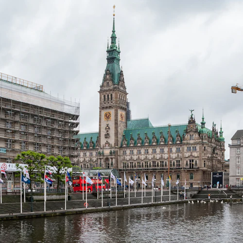 Historisk byggnad med ett klocktorn och gröna tak, ställningar på intilliggande byggnad, flaggor längs vattnet och en kran i bakgrunden.