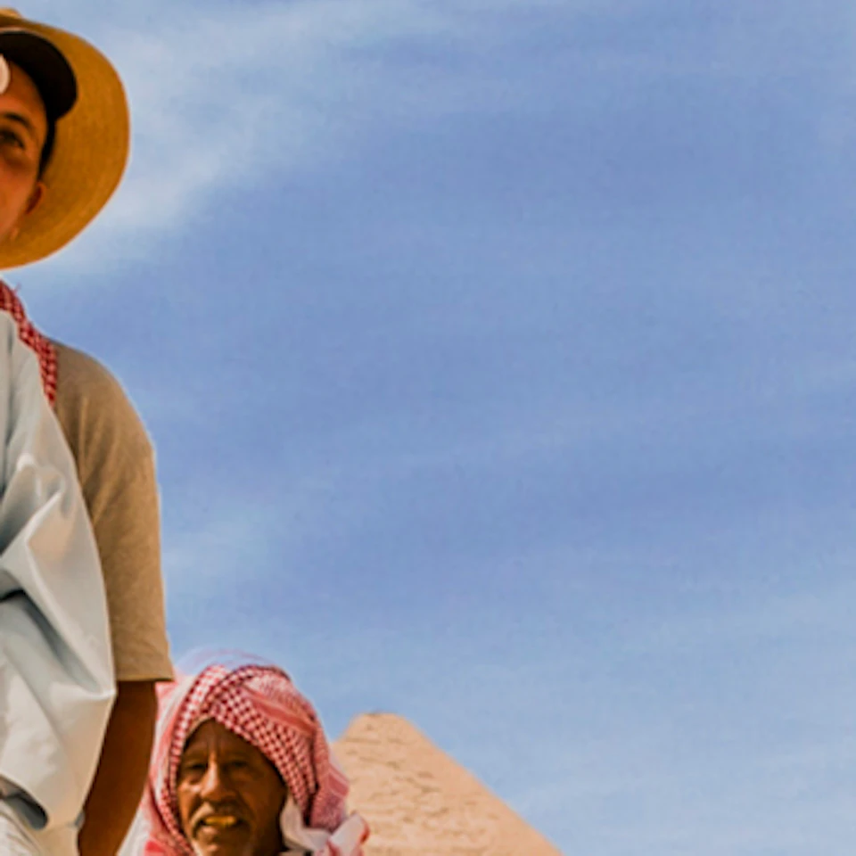 Tourists ride camels, adorned with colorful blankets, near the pyramids under a bright blue sky.
