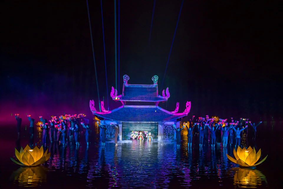 A theatrical performance on water with vibrant lighting, a traditional building backdrop, and performers in colorful attire.