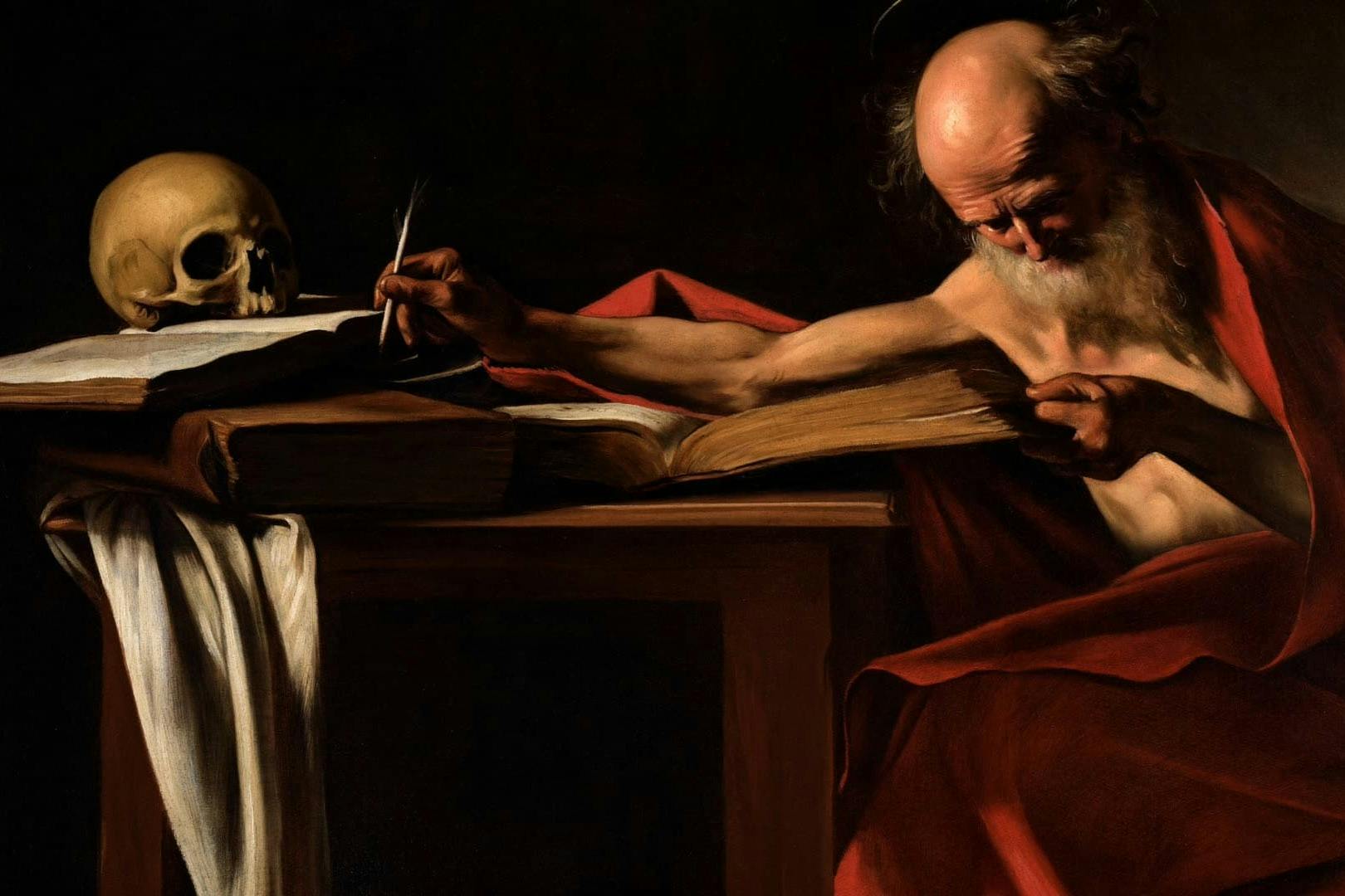 A hand holding a quill above an open book on a wooden desk, with a skull and a draped cloth visible on the desk.