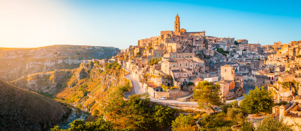 Historic hillside town with clustered stone buildings, a tall bell tower, winding roads, and lush vegetation under a clear blue sky.