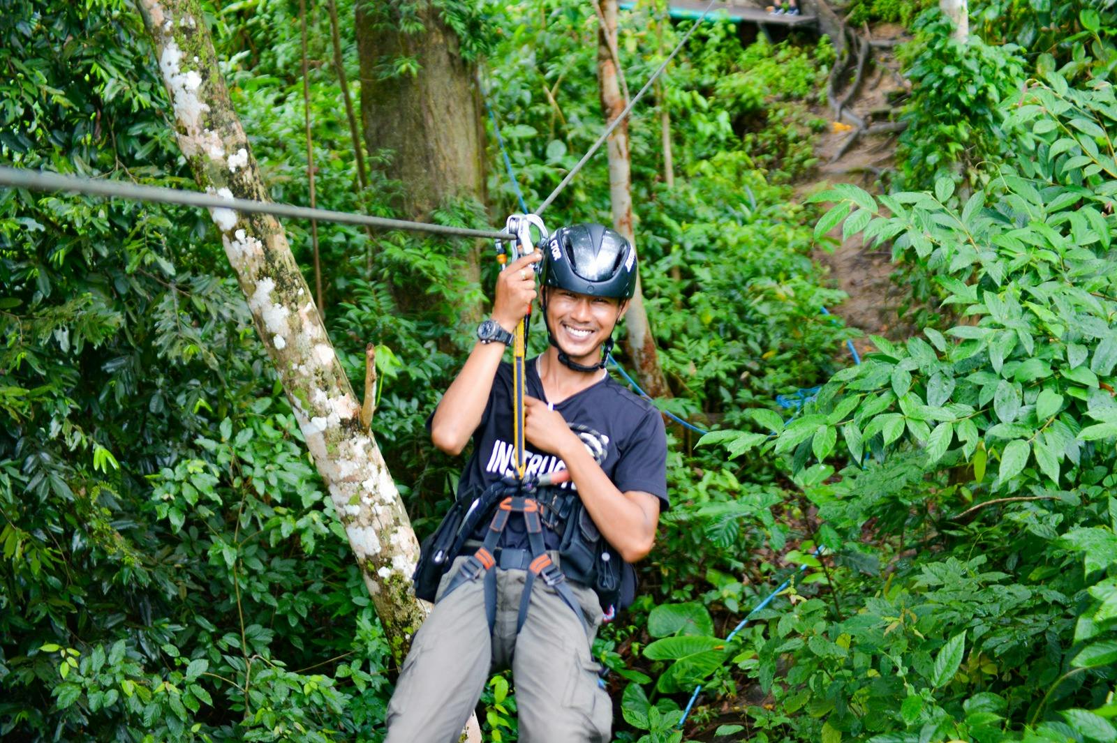 Persona sonriendo mientras se desliza en tirolina por un frondoso y verde bosque, con casco y arnés.