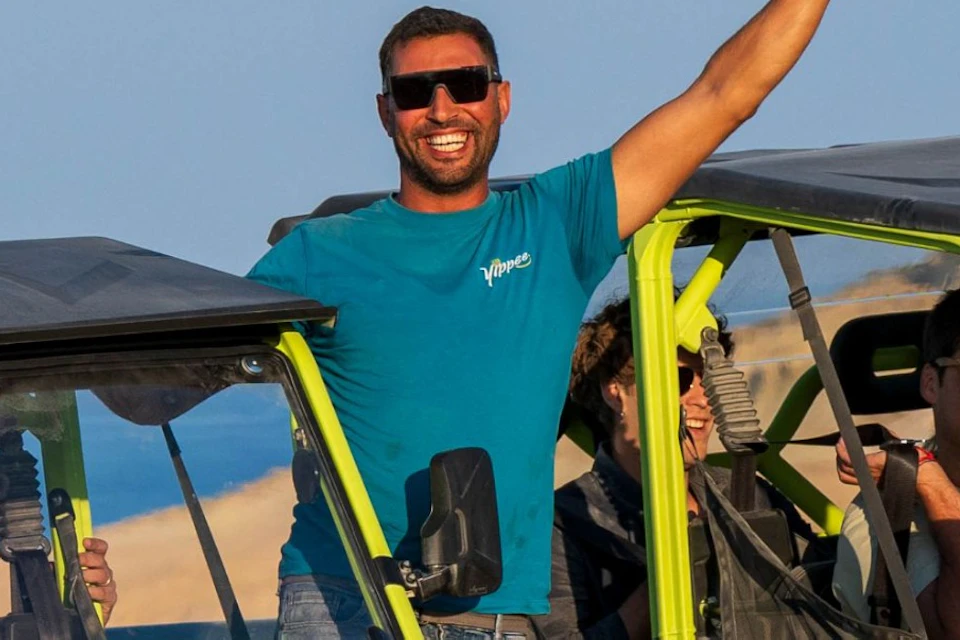 People in open-top utility vehicles on a sandy terrain, smiling and waving under a clear sky.