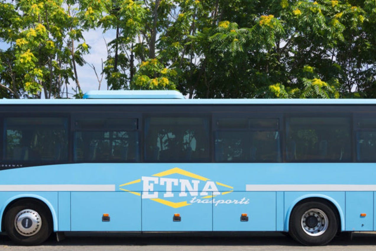 노란색 꽃이 핀 나무 앞에 'ETNA trasporti' 라고 표시된 파란색 버스가 주차되어 있습니다.