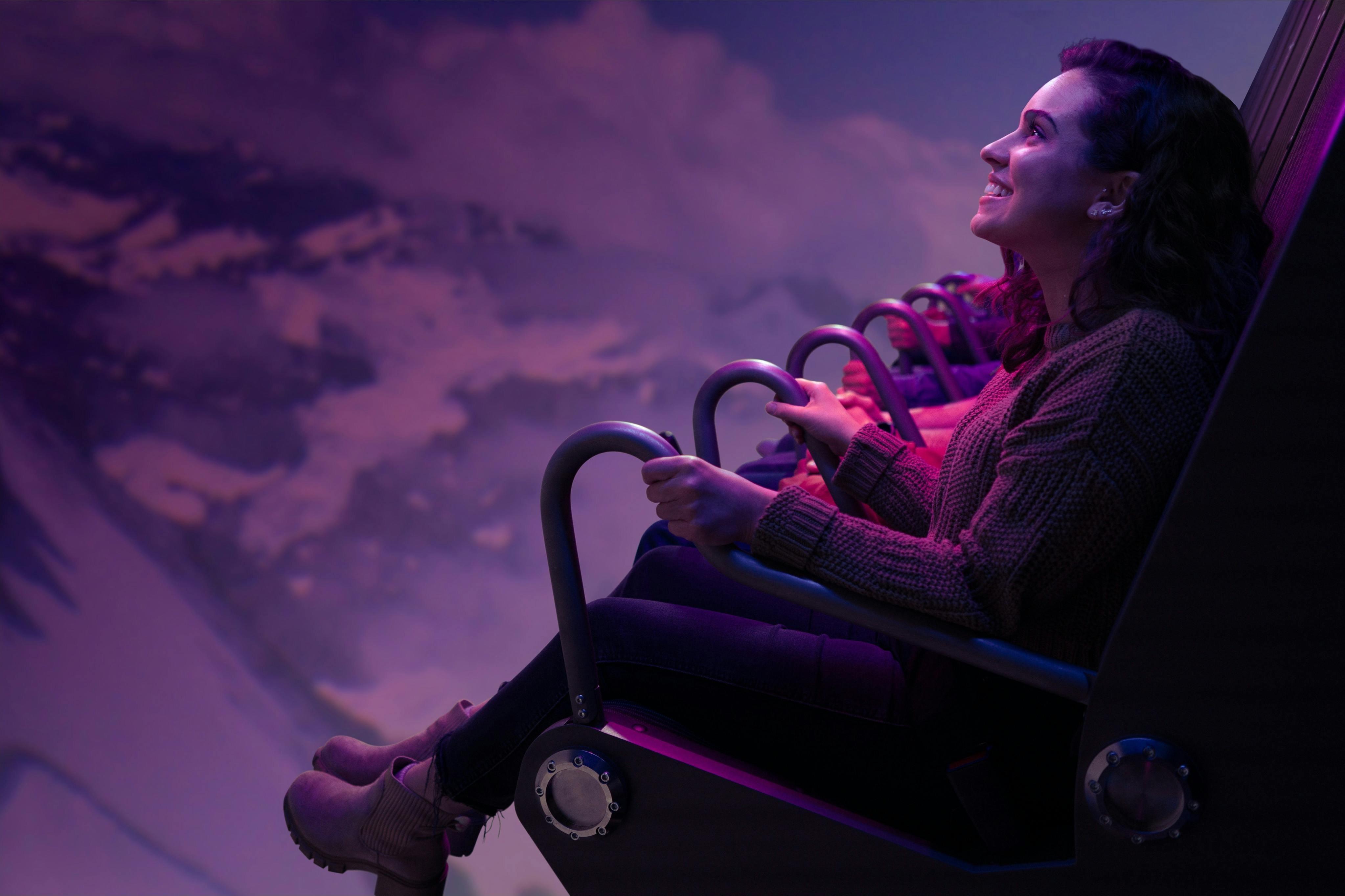 A person seated in an amusement ride, smiling, with a scenic backdrop illuminated by purple lighting.