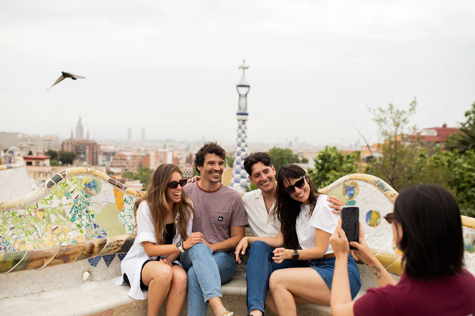 Four people seated on a colorful mosaic bench smiling, with a cityscape and decorative column in the background.