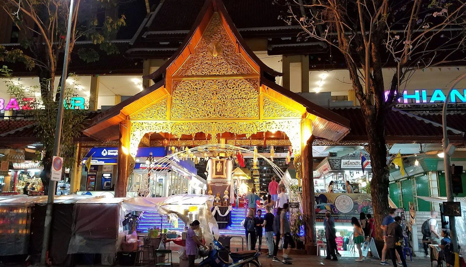 Ornate illuminated temple entrance with people nearby, surrounded by shops and decorations in an evening setting.